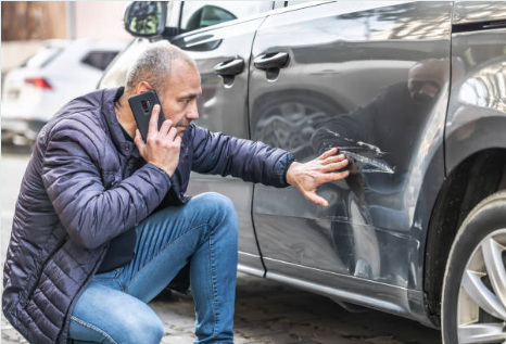 A man kneels beside a car, examining a scratch on the door while talking on his phone. He looks concerned, and wears a dark jacket and jeans.